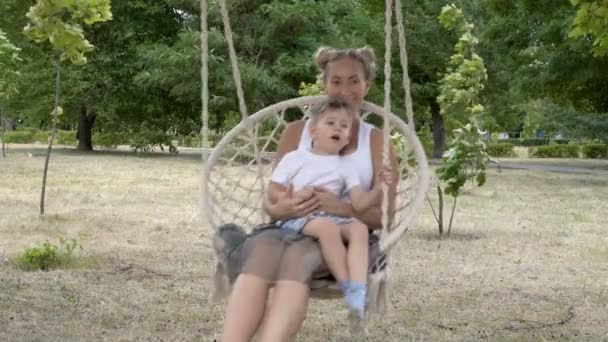 若いお母さんと赤ちゃんは、公園でぶら下がりブランコに座ってスイングして遊びます。子供は母親の膝の上に座り、晴れた日に緑の木々を背景に笑い、笑う。クローズ アップ。4k. — ストック動画