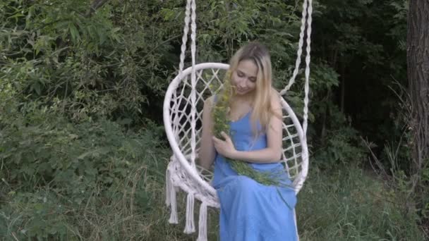 Una chica joven y sonriente se balancea en un columpio con un ramo de flores silvestres amarillas al atardecer de un día de verano al aire libre con un vestido azul. Ella sonríe y parece soñadora. 4K . — Vídeo de stock