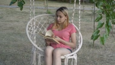Bir yaz gününün gün batımında bir şehir Parkı'nda beyaz asılı salıncak üzerinde otururken bir kitap ile yapraklı güzel bir genç kız. Close. 4k.