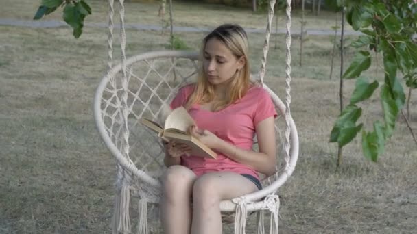 Ładna młoda dziewczyna przeglądając książkę siedząc na białym wiszącym huśtawce w parku miejskim o zachodzie słońca letniego dnia. Zbliżenie. 4K. — Wideo stockowe