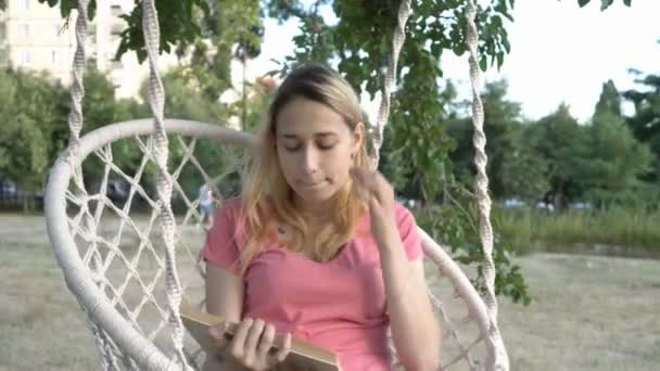 Крупный план молодой девушки, читающей книгу на качелях в парке на закате. Она выпрямляет волосы и качается на белых качелях в розовой футболке на фоне зеленых растений. Вид спереди. 4K . — стоковое видео