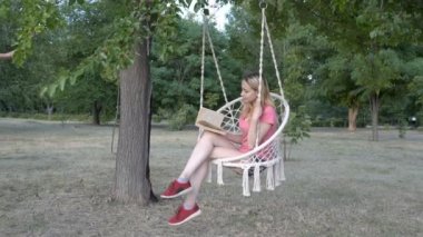 Güzel bir genç kız, sarışın, düşünceli bir yaz gününün gün batımında bir şehir Park'ta bir salıncak bir kitap okuma. O yeşil bitkilerin bir arka plan karşı pembe bir t-shirt beyaz bir salıncak üzerinde salıncak.