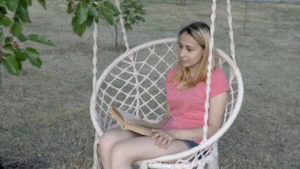 Uma menina loira bonita com um livro balança em um balanço em um parque da cidade no por do sol de um dia de verão. Ela sorri e olha para o lado. Close-up. 4K . — Vídeo de Stock