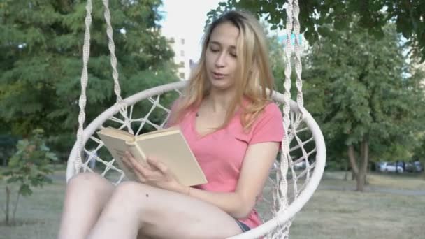 Una joven con el pelo desaliñado lee un libro y se balancea en un columpio hecho de cuerdas en un parque de la ciudad al atardecer, y sonríe. Primer plano. Vista frontal desde la parte inferior. 4K . — Vídeo de stock