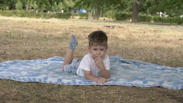 Un niño encantador está acostado boca abajo sobre la hierba en el parque de la ciudad al atardecer de un día de verano. Cuelga las piernas, mira a su alrededor y sonríe. 4K . — Vídeo de stock