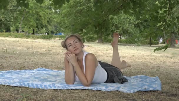 Bionda sexy con grandi seni sdraiata sull'erba nel parco al tramonto di una giornata estiva e penzola i piedi nudi. Alza lo sguardo pensieroso e sorride. Primo piano. 4K . — Video Stock
