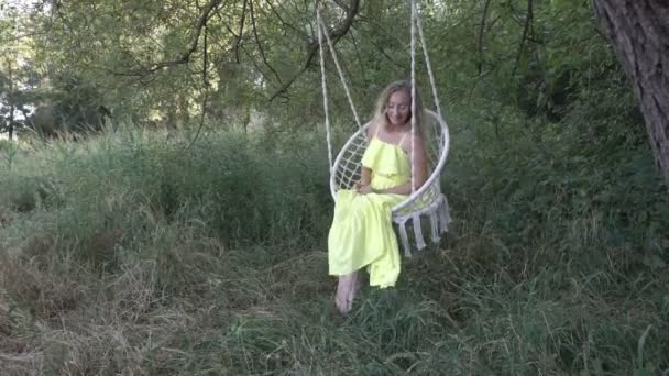 Sexy young blonde girl swinging on a swing outdoors in a yellow dress. She smiles, dangles her bare feet and poses for the camera at the sunset of a summer day against the green trees. Close-up. 4K. — Stock Video