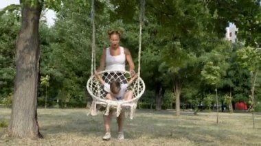 Mutlu bir genç anne parlak bir yaz gününde, Park'ta beyaz bir salıncak üzerinde küçük oğlu sallar. Gülerler ve yeşil ağaçlara karşı oynarlar. Aile mutluluğu kavramı. 4k