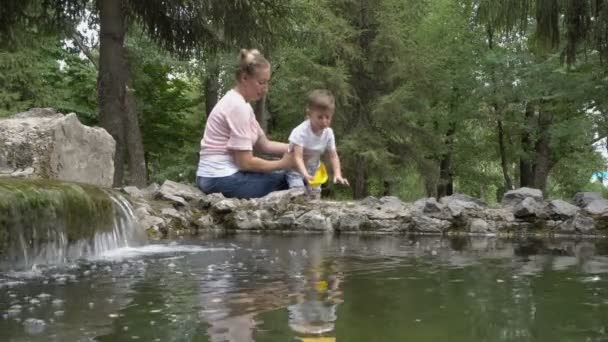 若い母親と小さな男の子は、市内の噴水で黄色の紙のボートを起動する準備をしています。赤ちゃんは笑い、喜びのためにジャンプします。家族のゲーム。概念。4k — ストック動画