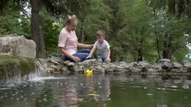 Bambino felice e mamma stanno lanciando una barca di carta gialla con una bandiera blu nella fontana della città. Il ragazzo ride e soffia sulla barca. Felicità familiare. Concetto. 4K — Video Stock