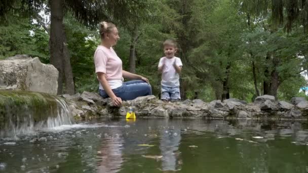 Een kleine jongen klappen zijn handen, springt voor vreugde en klappen op een gele papieren boot met een blauwe vlag in de fontein van de stad. Zijn moeder zit in de buurt, glimlachend en helpt het kind. Familie geluk. Concept. 4k — Stockvideo