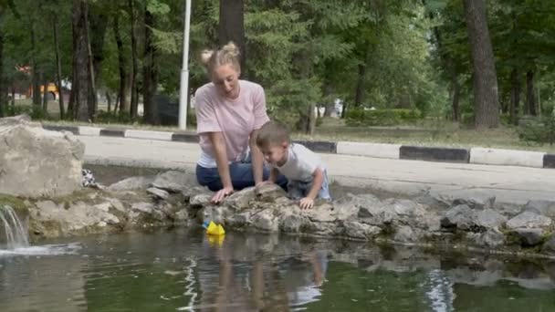 Een jongetje en zijn moeder spelen in het park aan het water met een gele papieren boot op een zomerdag. Baby en moeder om beurten zoenen elkaar. Concept. 4k — Stockvideo