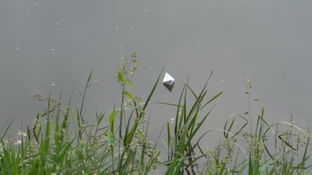 De wind drijft de White Paper Boat op het gladde oppervlak van het water. Uitzicht vanaf de kust door groene planten. Close-up. 4k — Stockvideo