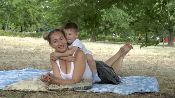 Счастливый ребенок сидит на спине своей матери, которая лежит на траве, смеется и обнимает свою материнскую шею. Мама и маленький мальчик играют на лужайке в городском парке в летний день. Крупный план. 4K — стоковое видео