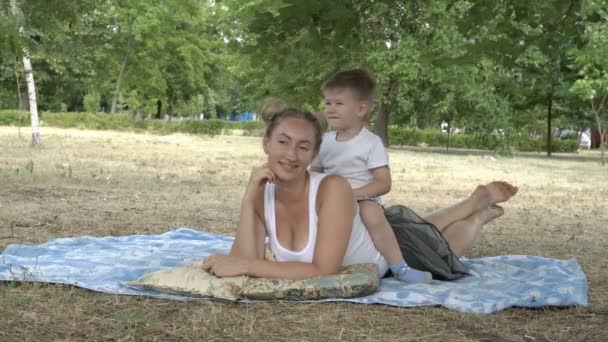 Joyeux maman et petit fils couchent, sourient et posent pour la caméra sur la pelouse dans le parc de la ville au coucher du soleil. Le bébé s'assoit sur le dos de sa mère et touche ses cheveux. Ils portent des chemises blanches. 4K — Video