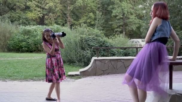 女摄影师在日落时分在公园里，在户外拍摄一个年轻女孩模型。她拿着一台带长焦镜头的照相机。总体视图。4k — 图库视频影像