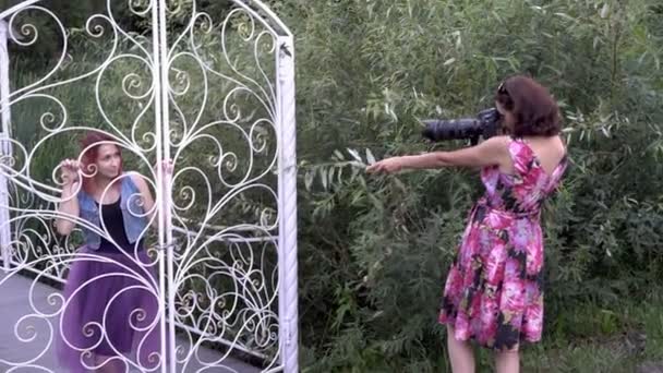Mãe-fotógrafa leva uma câmera sua filha adulta ao ar livre, no Parque, através dos ramos verdes. Ela está segurando uma câmera com uma lente telefoto. Vista lateral. 4K — Vídeo de Stock