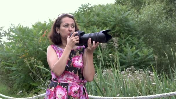 Ženská fotografka při práci venku. Drží fotoaparát s teleobjektiv, fotí obrázky a radí někomu za scénami v parku při západu slunce letního dne. 4k