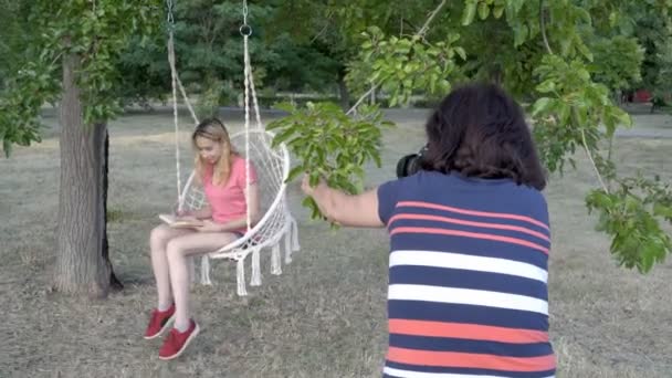 A young blonde girl posing for a photographer on a rope swing, with a book in her hands, in the Park at the sunset of a summer day. Close up. View from behind the photographer. 4K — Stock Video