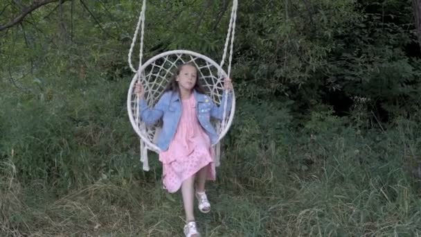 En söt bebis, en liten flicka, svänga på en vit hängande Swing i naturen på en solig dag. Hon ler och leker utomhus i en rosa klänning och jeansjacka. Närbild. 4K. — Stockvideo
