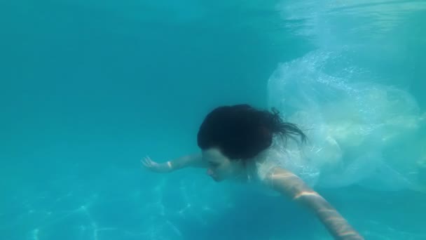 Une mariée extraordinaire dans une robe blanche nage lentement sous l'eau dans la piscine près du fond sur un fond bleu par une journée ensoleillée. Au ralenti. Gros plan. 4K . — Video