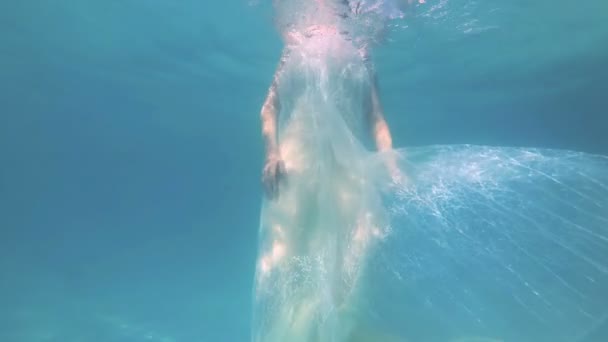 Uma noiva bonita em um vestido branco mergulha e posa debaixo d 'água na piscina na câmera com o cabelo para baixo em um fundo azul em um dia ensolarado. Movimento lento. Close-up. 4K . — Vídeo de Stock