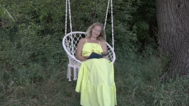 Очаровательная блондинка читает книгу, сидя на висячих качелях на природе в летний день. Она улыбается в желтом сарафане с синей книгой в руках на белых качелях. Крупный план. 4K . — стоковое видео