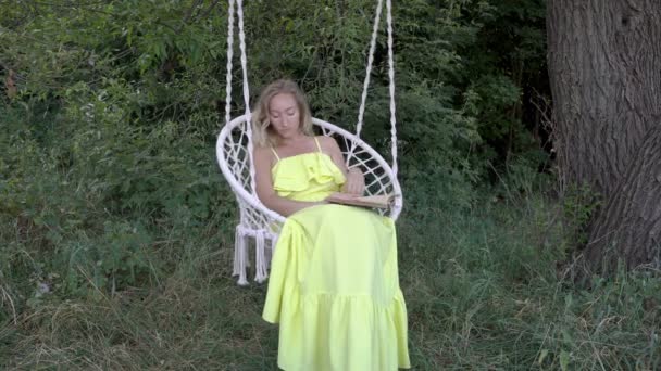 Piękna Blondynka siedzi na huśtawce w żółtej sukni i czytanie książki w przyrodzie w słoneczny dzień. Film koncepcyjny. Portret mody. Zbliżenie. 4K. — Wideo stockowe