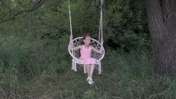 Zamyšlavá holčička se pokouší otočit na zavěšenou houpačku, která visí ze stromu v parku na pozadí zelených keřů. Dítě v růžových šatech na bílém houpačce. Close-up. 4k. — Stock video