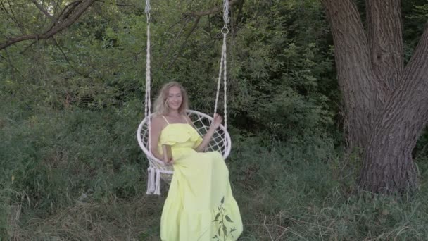 Glad blond flicka på en hängande Swing i en gul klänning i naturen på en solig dag. Hon håller en bok, rätar håret, skrattar provokativt och spelar på den vita repsvingen. RAW-video. 4K. — Stockvideo
