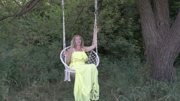 Una joven rubia sexy en un columpio se está divirtiendo jugando y colgando sus pies desnudos mientras está sentada en un vestido amarillo en la naturaleza en un día soleado. Vídeo crudo. 4K . — Vídeo de stock