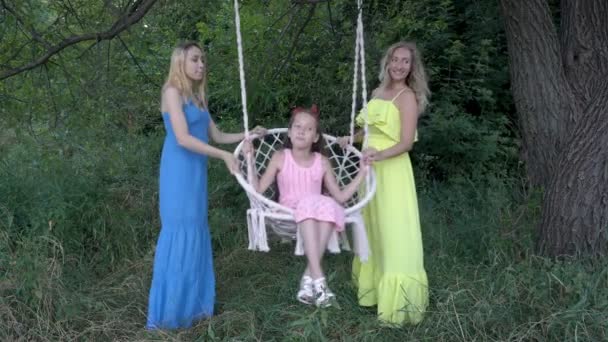 Två söta flickor skakar en liten flicka i en rosa klänning på en vit Swing utomhus på en solig dag. De ler och poserar i färgglada klänningar. Mode porträtt. Närbild. 4K. — Stockvideo