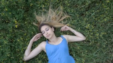 Üzgün bir genç kız kollarını uzanmış ve yukarı bakıyor yeşil çim üzerinde parkta sırt üstü yatıyor. Sarı kır çiçekleri ile çevrili mavi bir elbise giyiyor. Moda portresi. 4k.