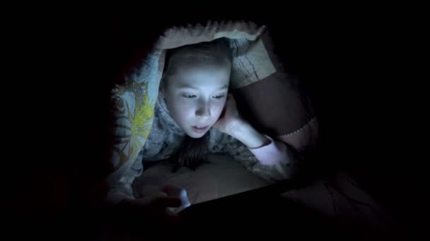 Uma menina brincando em um tablet à noite em uma cama debaixo de um cobertor. Vídeo conceitual. Close-up. Vídeo cru. 4K . — Vídeo de Stock
