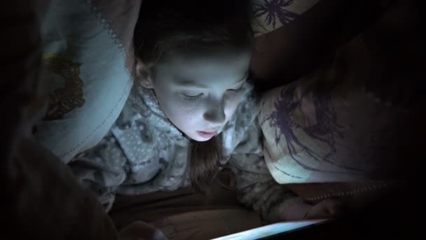 Ребенок, маленькая девочка, прятался под одеялом на кровати ночью и играл на планшете в темноте. Концепция видео. Интернет-зависимость. Крупный план лица. Сырое видео. 4K . — стоковое видео