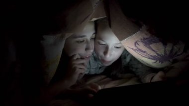 İki gülümseyen küçük kız tablette fotoğraf izliyor, geceleri karanlıkta yatağın üzerinde bir battaniyenin altında yatıyorlar. Konsept video. İnternet bağımlılığı. Çocukların yüzlerini kapatın. Ham video. 4k.