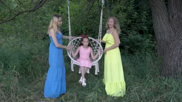 Tres hermanas de diferentes edades juegan y se divierten en la naturaleza en un día soleado con vestidos coloridos. Ellos rockean a la hija más joven en el columpio y sonríen. Retrato de moda. Primer plano. 4K . — Vídeo de stock