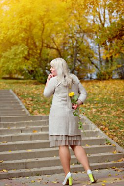 Sevimli sarışın büyük boy standları ve bir taş merdiven karşı, Park güneşli bir sonbahar gününde arkasında sarı bir gül tutar. Düşünceli bir şekilde uzaklara bakıyor. Kavramsal fotoğrafçılık. Arka görüş