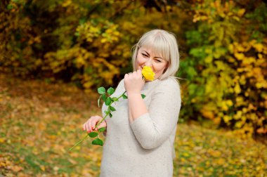 Büyüleyici bir orta yaşlı sarışın kadın duruyor ve güneşli bir sonbahar gününde bir Parkta poz veriyor. Sarı bir gülü kokluyor ve gülümsüyor. Kavramsal fotoğrafçılık.