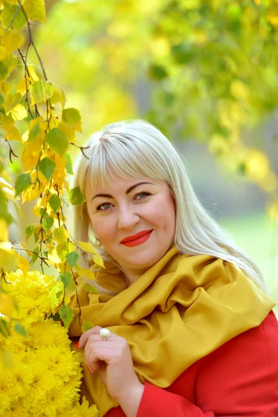 Retrato de uma linda loira de meia-idade ao ar livre. Ela fica perto de uma bétula com folhas amarelas em um manto vermelho com um buquê de flores amarelas, olha para a câmera e sorri . — Fotografia de Stock