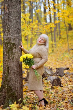 Narin örme elbiseli bir sarışın ormanda güneşli bir sonbahar gününde poz veriyor. Rüya gibi görünüyor. Dudaklarındaki kırmızı ruj. Kadın boyutu artı xxl.