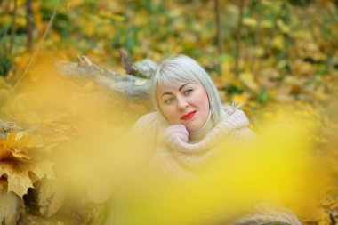 Parlak sonbahar arka planda Muhteşem sarışın ormanda nazik bir jumper poz yaprakları, günlükleri eğilerek ve düşünceli uzağa bakarak gülümseyerek. Kadın artı boyutu xxl. Yatay görünüm
