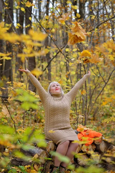 Lächeln Blondine wirft im Wald Herbstlaub an einem sonnigen Tag. ist glücklich und blickt auf. Frauen plus size xxl. vertikale Ansicht. — Stockfoto