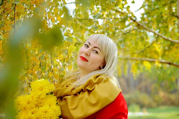 Plus size blonde woman with a bouquet of yellow flowers outdoors. She stands against a background of trees with yellow leaves in a red raincoat, looking at the camera and smiling. Fashion portrait
