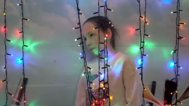 Malá holčička, která si zahrává s vánočními světly stojící za nimi u zdi domu. Usmívá se a dívá se na kameru skrz blikající světla. Close-up. 4k. — Stock video