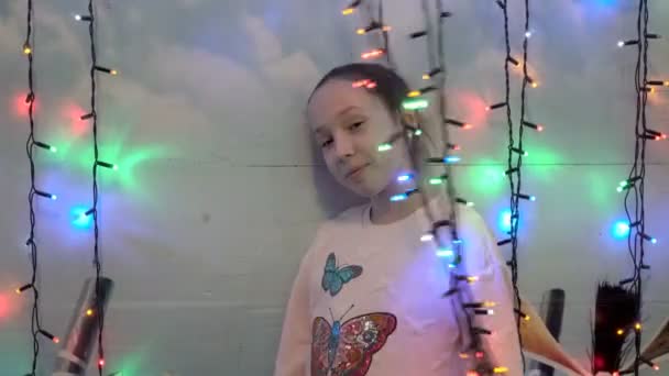 Petite fille souriante jouant avec des guirlandes suspendues colorées se tenant derrière eux près du mur de la maison. Elle sourit et regarde la caméra à travers les lumières clignotantes. Gros plan. 4K . — Video