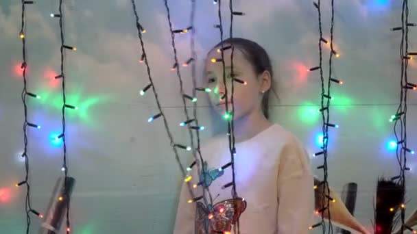 Un enfant, une petite fille, se tient derrière des guirlandes colorées près du mur de la maison. Elle touche ses mains et joue avec les lumières clignotantes. Gros plan. 4K . — Video