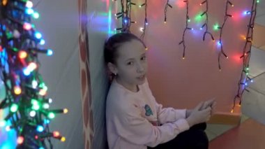 Küçük bir kız renkli Noel çelenk arka planda duvara yakın oturan, kameraya bakarak ve gülümseyerek. Portre. Close. 4k.