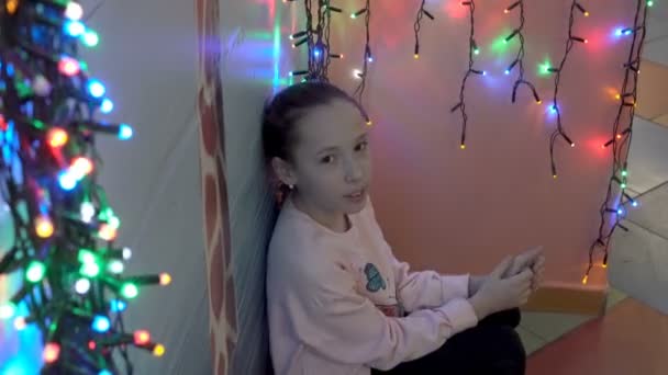 Ein kleines Mädchen, das neben der Wand auf dem Hintergrund bunter Weihnachtsgirlanden sitzt, in die Kamera blickt und lächelt. Porträt. Nahaufnahme. 4k. — Stockvideo