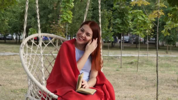 Linda joven descansando en un columpio blanco en el parque de la ciudad. Lee un libro y se ríe provocativamente en un día soleado de otoño. Está cubierta con una manta roja. Retrato. Primer plano. 4K . — Vídeos de Stock